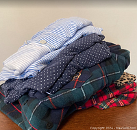 Stack of four folded pajamas sets showing blue/white stripes, navy polka dots, green plaid fleece, and red plaid with leopard print trim.