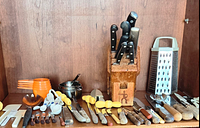 Wide shot of kitchen utensils including wooden knife block, stainless steel grater, orange measuring cups, yellow peelers, and various kitchen gadgets laid out on wooden surface.