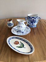 Four decorative pieces arranged on a wooden surface, showing the full lot including the Delft oval plate, two-handled Delft pot, Royal Copenhagen pitcher with plate, and the small unidentified pitcher.