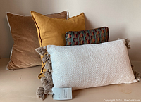 Four throw pillows arranged side by side on a surface showing fabric textures and colors. Tags visible on the white woven pillow.