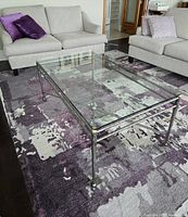 Angled view of the rectangular steel and glass coffee table placed on a purple patterned rug between two sofas.