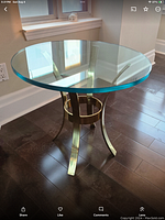 Full view of round bevelled glass top and polished brass curved leg base on hardwood floor near window