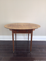 Front view of the oval drop leaf side table showing tapered legs on casters and dovetailed drawer below the tabletop.