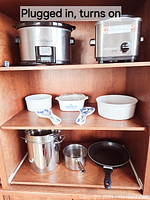 Front view of small stainless steel Cuisinart slow cooker, Cuisinart rice cooker, CorningWare dishes with blue cornflower pattern, and assorted stainless steel pots and pans on wooden shelving