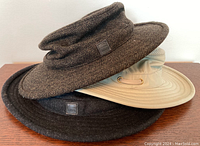Three Tilley hats stacked: brown herringbone wool TEC, charcoal wool TEC, beige/khaki Airflo sun hat