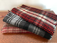 Four folded plaid scarves stacked showing distinct colors and patterns, with fringed edges visible, placed on a wooden surface.