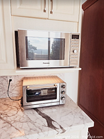 Panasonic microwave oven placed on a shelf above a marble countertop. Below on the counter is a Cuisinart toaster oven.