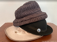 Three hats stacked: black wool felt Tilley Oslo Hat on bottom, beige Tilley Airflo hat in middle, brown Lizzy By Design quilted bucket hat on top.