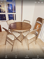 Photo showing the vintage round folding table with dark brown upholstered top and four matching beige upholstered folding chairs around it, plus two additional metal folding chairs stacked nearby.