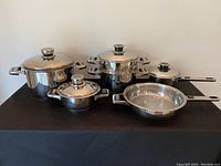 Cookware set displayed showing multiple stainless steel pots with matching lids and a frying pan on a black surface.