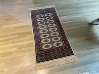 Full view of Bokhara design runner rug laid flat on light wood floor showing red and blue bordered geometric designs and cream fringe on one end.