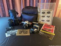 Wide view of Nikon FG film camera with attached 50mm f/1.8 lens, zoom lens, teleconverter, flash unit, multiple cases, instruction manuals, and large camera bag on black cloth background