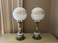 Pair of VV Vianne lamps with white glass globes decorated with gold scrollwork and cherub brass bases on marble