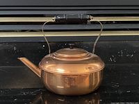 Copper kettle with black handle shown from side angle against dark background, highlighting the full profile and spout design.