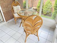 Two rattan fan-back armchairs and round glass-top table arranged in a sunroom, angled view showing overall set.