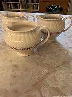 Three bone china jugs with handles, cream colored, with textured surface and floral band around midsection, shown on marble-like surface.