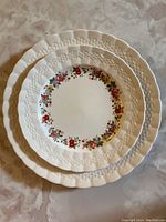 Stack of round bone china plates with scalloped, embossed rims and floral wreath design in center, varying sizes visible.
