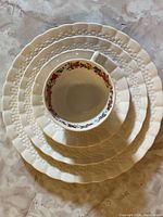 Stacked view of white plates and a cup with saucer showing embossed rim and floral wreath interior on cup.