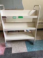 Front view of cream colored metal rolling book cart with three shelves and casters.