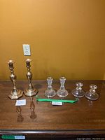 Photo showing the full set of six assorted candlestick holders: 2 gold-tone metal, 2 clear glass with beaded designs, 2 clear glass with etched/floral designs, all displayed on a wood surface against a yellow wall.