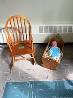 Wooden child's rocking chair and wooden doll cradle with baby doll and blanket placed on floor near heating vent.