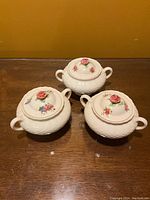 Three white vintage china sugar bowls with lids on a wooden surface under yellow-orange background. Each bowl has two handles and floral decoration on the lids. One lid has a notable chip.