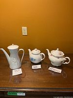 Group photo of the coffee pot, Corelle stoneware teapot, and china teapot on a wooden surface with informational labels.