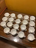 Full view of all 19 white ceramic Corning mugs arranged on a wooden surface, showing their shape and handle style.