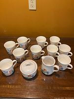 Full view of the 10 mugs, sugar bowl with lid, and creamer all featuring the floral pattern on white Corning Ware.