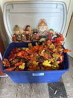 Photo showing five fall scarecrow figurines standing behind a large blue storage bin filled with artificial fall leaves and garlands.