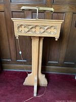 Full view of the oak lectern against wooden panel background, showing carved front decorative panel and brass reading light attached on top.