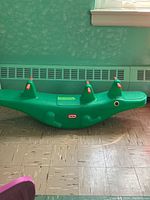 Side view of green plastic alligator toddler riding toy with three orange handle grips and rounded teeter base on tiled floor against teal wall