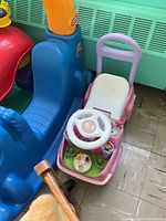 Pink and white ride-on toy with steering wheel and activity toys on base, large blue ride-on animal toy and part of red toy visible