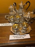 Full view of six-piece silverplate tea set arranged on a wooden surface with two trays, showing tarnish and reflective surfaces.