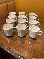 Front angled view showing the group of 30 white mugs arranged in rows on wooden surface.