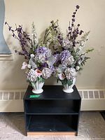 Two floral arrangements with purple, lavender, white, and pink artificial flowers in white plastic vases placed on black wooden stand with two shelves.