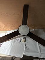 Top view of ceiling fan showing three dark wood tone blades, central textured glass light fixture, and remote control on white cloths.