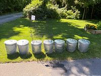 Seven half-size galvanized metal cans with lids and an iron planter frame placed on grass outdoors.