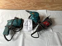 Overview photo shows three power tools: one red drill, one teal drill, and one teal palm sander, all corded and used.