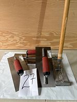 Three drywall trowels arranged on white surface with wood background, showing red handles and metal blades.