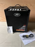 Front view of the Peavey MAX 110 amplifier with manual and power cord placed next to it on the floor.