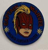 Front view of a round Captain Marvel collector pin with a blue background, featuring the character's masked face and text around the edge.