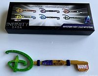 Photo of the Marvel Studios The Infinity Saga collector mystery key displayed outside of its packaging. The key has a green glitter Disney D logo, gold shaft with "I AM INEVITABLE", and Marvel logo on the handle.