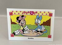 Front of trading card showing Minnie and Daisy playing marbles with colorful illustrated scene and decorative border.
