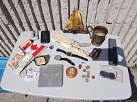 Entire lot arranged on a white outdoor table showing all items spread out including watches, currency, cutters, stamps, metal cup, and boxes.
