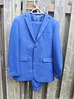 Full view of blue men's suit jacket and folded pants on hanger, displayed against wooden fence.