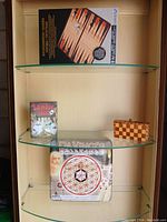 4 board games displayed on glass shelving including Da Vinci's Challenge, Survival Juniors, backgammon game, and a wooden folding checkered board.