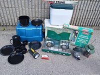 Lot setup showing all items: black camping cookware, Coleman propane stove with box, two vintage lanterns, two coolers, flashlight, and Coleman high stand.