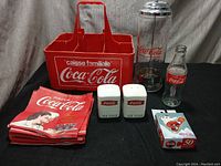 Red plastic Coca-Cola crate, glass bottles, salt and pepper shakers, napkins, and playing cards laid out on black fabric.