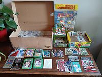 Image showing a large open box filled with baseball cards, a yellow box with additional baseball cards, GI Joe card packs, and loose cards arranged on the table.